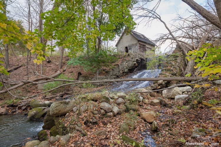 An abandoned pumphouse sits beside a waterfall in the woods near the railroad tracks.